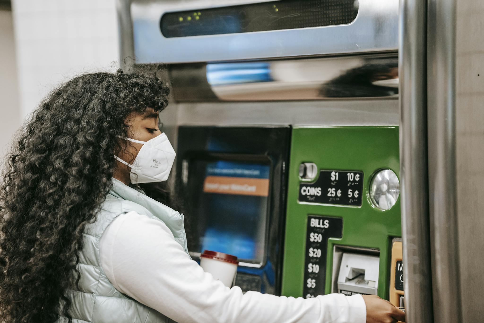 A woman wearing a face mask uses a ticket machine for purchasing fare in a public setting, reflecting safety during pandemic times.